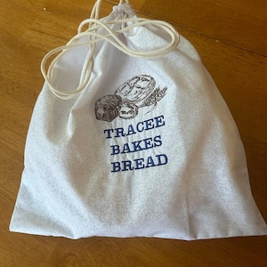 May include: White cotton drawstring bag with a blue and brown embroidered design of bread loaves and the text "Tracee Bakes Bread".