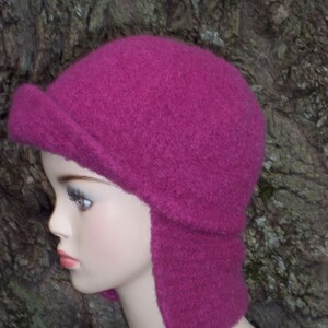 May include: A pink knitted hat with ear flaps. The hat is made of a soft, fuzzy material and has a simple design.