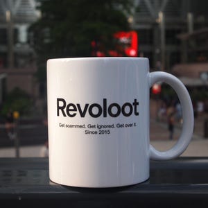 Op de afbeelding: Witte keramische mok met het woord "Revoloot" in zwart, samen met de zinnen "Get scammed. Get ignored. Get over it." en "Since 2015." De mok heeft een gebogen handvat. De achtergrond is wazig, met een buitenscène.