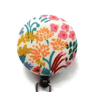 May include: A circular badge reel featuring a cream-colored fabric cover with a vibrant floral pattern. The design includes stylized flowers and leaves in shades of pink, orange, yellow, and teal, attached to a black plastic base with a metal clip.