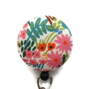 May include: A circular badge reel featuring a white fabric cover with a colorful floral pattern. The design includes pink daisies, small orange flowers, and teal green leaves printed on a textured textile surface, attached to a black plastic base.