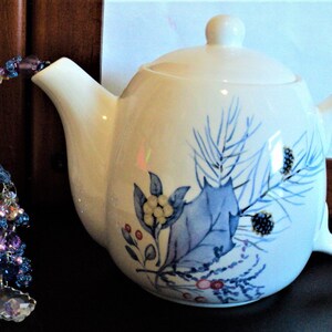 Puede incluir: Tetera de cerámica blanca con un diseño floral azul y morado. La tetera tiene una tapa y un asa. Un borla de cuentas cuelga del pico.