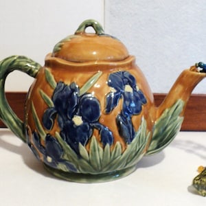 May include: A ceramic teapot with a lid, featuring a brown and orange glaze. The teapot is decorated with blue iris flowers and green leaves. A beaded ornament hangs from the spout. The handle and spout are green.