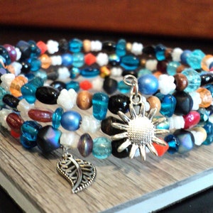 May include: Multi-strand bracelet with blue, red, orange, white, and black beads. The bracelet has two charms: a silver sunflower and a silver leaf.