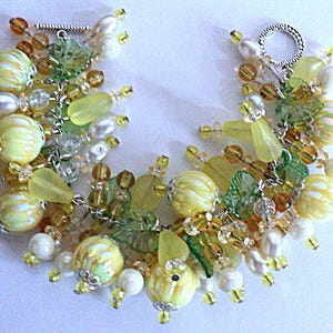 May include: A handcrafted bracelet featuring a mix of yellow, green, and clear beads. The bracelet includes large, round, yellow beads with white stripes, smaller yellow and clear beads, and faux pearls. The clasp is silver-toned.