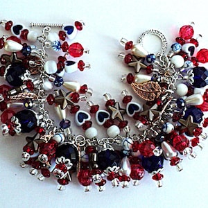Op de afbeelding: Een patriottische bedelarmband met rode, witte en blauwe kralen, sterren en hartvormige bedels. De armband heeft een verscheidenheid aan texturen en vormen, waaronder facetkralen, parels en metalen accenten. De armband heeft een tuimelsluiting.