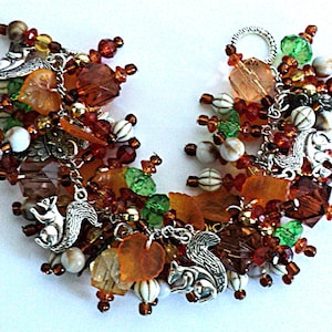 May include: A charm bracelet featuring silver-toned squirrel charms interspersed with amber, brown, green, and white beads. The bracelet includes various faceted crystal beads, leaf-shaped charms, and round striped beads attached to a silver chain.