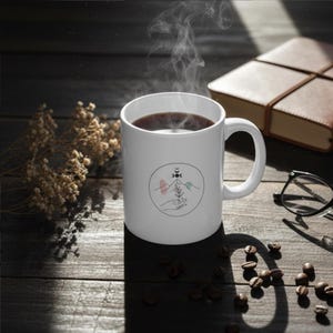 May include: A white ceramic coffee mug filled with dark liquid, possibly coffee, with steam rising. The mug features a circular design with mountains, a crescent moon, and a hand holding a plant. The mug is on a wooden surface with coffee beans, a book, and glasses.