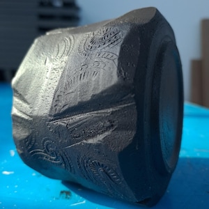 Peut inclure: Une tasse en céramique gris foncé, facettée, avec des motifs floraux et géométriques gravés. La tasse a une forme cylindrique avec une surface légèrement texturée. Le motif est sculpté dans la surface, créant un contraste tactile et visuel.