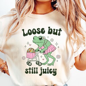 May include: A white t-shirt with a green frog wearing a pink tutu and pushing a disco ball in a stroller. The text "Loose but still juicy" is printed on the shirt.