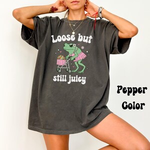 May include: A dark gray t-shirt with a graphic of a frog wearing a pink tutu and pushing a baby carriage. The text on the shirt reads "Loose but still juicy" and "Pepper Color".