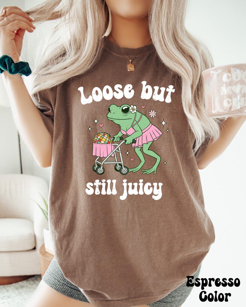 May include: A brown t-shirt with the text 'Loose but still juicy' in white. A cartoon frog wearing a pink bikini and a pink tutu is pushing a baby stroller with a disco ball inside. The frog is green with black eyes and a pink flower on its head.