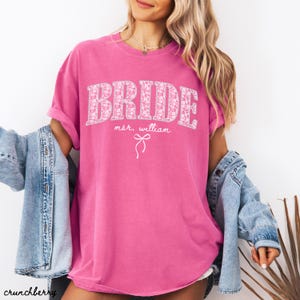 May include: A bright pink t-shirt with the word "BRIDE" in large, floral-patterned letters. Below the word "BRIDE" is the text "mr. william" and a bow graphic. The shirt has rolled-up sleeves.