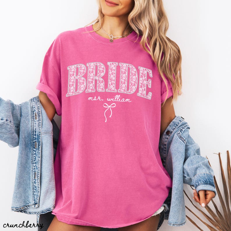 May include: A bright pink t-shirt with the word "BRIDE" in large, floral-patterned letters. Below the word "BRIDE" is the text "mr. william" and a bow graphic. The shirt has rolled-up sleeves.