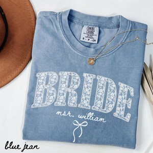 May include: A blue jean Comfort Colors t-shirt with the word "BRIDE" in white floral print. The text "msr. william" and a bow graphic are below the word "BRIDE". A gold necklace with a pendant is on the shirt.