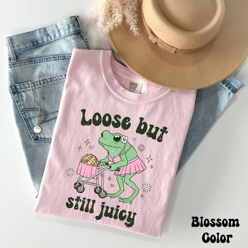 May include: Pink t-shirt with a green frog pushing a stroller with a disco ball. The text on the shirt reads "Loose but still juicy".