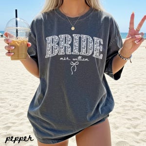 May include: A dark gray t-shirt with the word "BRIDE" in white floral print. Below the word "BRIDE" is the text "msr. william" and a small bow graphic. The word "pepper" is printed in the bottom left corner.