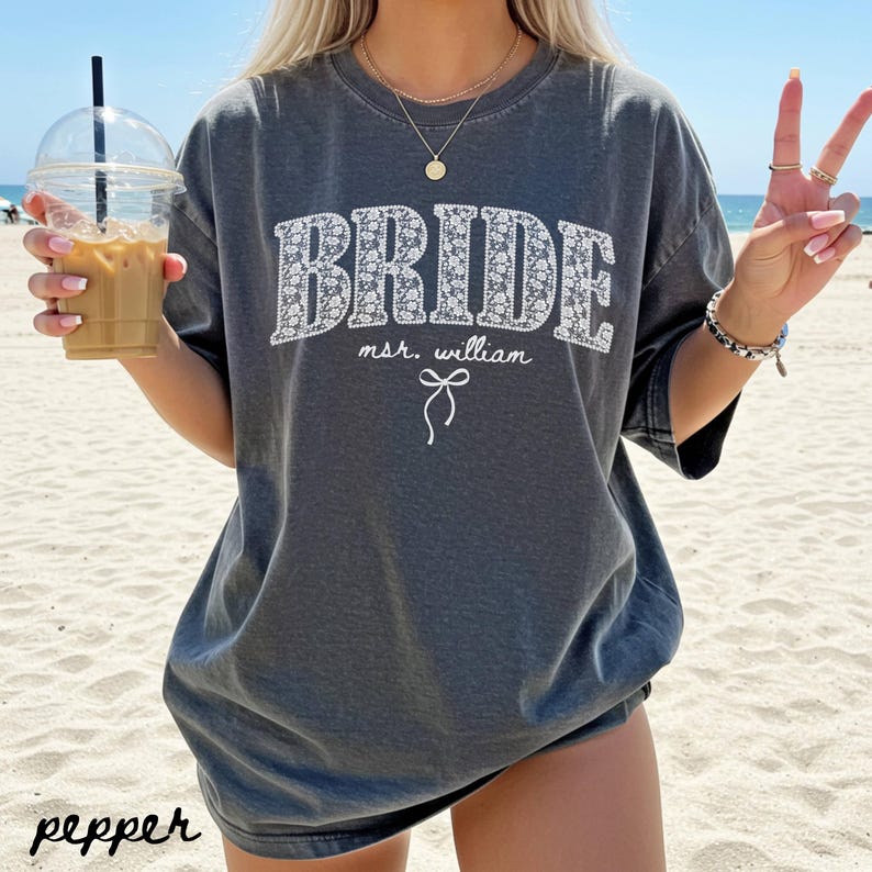 May include: A dark gray t-shirt with the word "BRIDE" in white floral print. Below the word "BRIDE" is the text "msr. william" and a small bow graphic. The word "pepper" is printed in the bottom left corner.