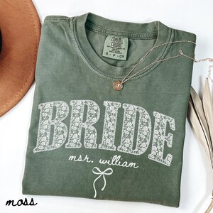 May include: A moss green t-shirt with the word "BRIDE" in white floral print. Below the word "BRIDE" is the text "msr. william" and a white bow graphic. A gold necklace with a pendant is on the shirt.