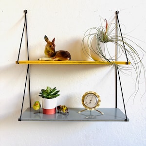 May include: A two-tiered hanging shelf with black supports. The top shelf is yellow, holding a ceramic deer figurine and a white vase with a plant. The bottom shelf is grey, with a potted succulent, small bird figurines, and a brass clock.