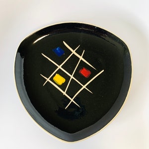 May include: A black, triangular ceramic plate with a white rim. The plate features a white grid design with three colored squares: blue, yellow, and red. The plate is likely for serving food.