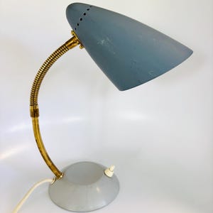 May include: A desk lamp with a conical blue metal shade featuring a row of small ventilation holes. The lamp has a flexible brass-toned gooseneck arm attached to a circular light gray base with a white toggle switch and a white power cord.