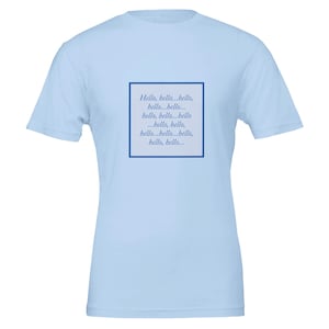 Puede incluir: Una camiseta de manga corta de color azul claro con un gráfico cuadrado central con un borde azul oscuro. Dentro del cuadrado, la palabra "Hello" se repite varias veces en una fuente serif cursiva de color azul oscuro, creando un patrón de texto.