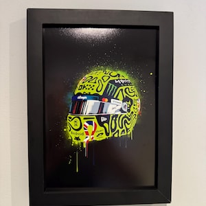 May include: Framed artwork of a neon yellow racing helmet with a black and white abstract design. The helmet features sponsor logos and the Union Jack flag. The background is black with green and yellow paint splatters.