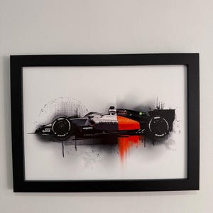 Può includere: Stampa artistica incorniciata di una macchina da corsa di Formula 1 in nero, arancione e bianco. L'auto è raffigurata in uno stile dinamico e artistico con effetti di schizzi di vernice. La cornice è nera.