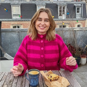 Puede incluir: Una mujer con un cárdigan a rayas rosa y blanco se sienta en una mesa de madera con una taza de café y una galleta de chocolate.