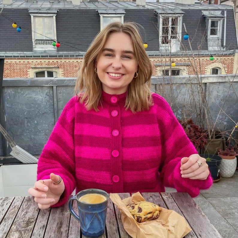 以下が含まれることがあります： ピンクと白のストライプのカーディガンを着た女性が、木のテーブルに座って、コーヒーとチョコレートチップクッキーを手にしています。