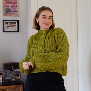 Op de afbeelding: Een groene, dikgebreide cardigan met een hoge kraag en grote, ronde knopen. De mouwen zijn lang en volumineus. De cardigan heeft een levendige groene tint en het breipatroon is getextureerd.