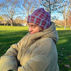 Może przedstawiać: Ręcznie robiona czapka z szydełka we wzór kratki w kolorze bordowym i jasnoszarym. Osoba ma na sobie oliwkową kurtkę puchową. Zdjęcie zrobiono na zewnątrz w słoneczny dzień.