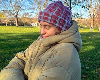 May include: A handmade, crocheted beanie in a grid pattern of burgundy and light gray. The hat is worn outdoors on a sunny day. The person is wearing a large, olive green puffer jacket.
