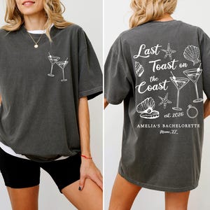K&ouml;nnte beinhalten: Dunkelgraues T-Shirt mit wei&szlig;er Grafik von zwei Martinigl&auml;sern auf der Vorderseite. Auf der R&uuml;ckseite steht der Text "Last Toast on the Coast" mit Muscheln, Seesternen, Martinigl&auml;sern und einem Ring. Mit "Amelia's Bachelorette" und "Miami, FL".