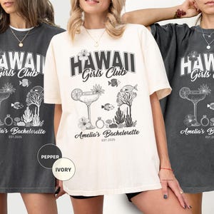 K&ouml;nnte beinhalten: Drei T-Shirts in Pfeffer- und Elfenbeinfarben mit einem grafischen Design. Das Design enth&auml;lt den Text "HAWAII Girls Club" &uuml;ber einem Cocktail, Fischen und floralen Elementen. Darunter steht "Amelia's Bachelorette EST. 2025".
