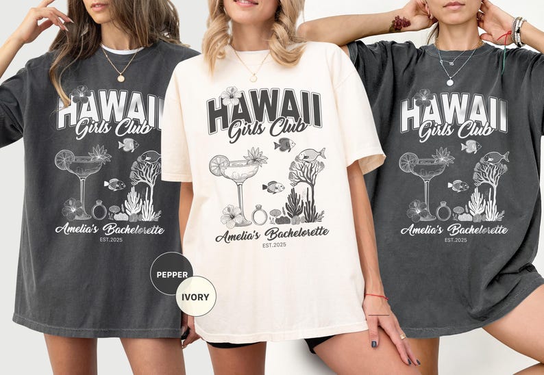 K&ouml;nnte beinhalten: Drei T-Shirts in Pfeffer- und Elfenbeinfarben mit einem grafischen Design. Das Design enth&auml;lt den Text "HAWAII Girls Club" &uuml;ber einem Cocktail, Fischen und floralen Elementen. Darunter steht "Amelia's Bachelorette EST. 2025".