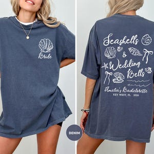 K&ouml;nnte beinhalten: Denimblaues T-Shirt mit wei&szlig;er Muschel und "Bride"-Text auf der Vorderseite. Die R&uuml;ckseite zeigt den Text "Seashells & Wedding Bells" und Illustrationen. Amelia's Bachelorette, Key West, FL, 2026.