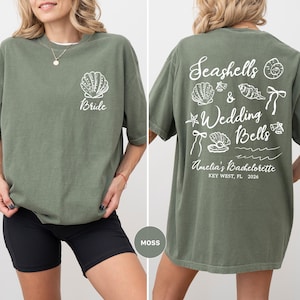 K&ouml;nnte beinhalten: Moosgr&uuml;nes T-Shirt mit einem Muschelmuster und dem Wort "Bride" auf der Vorderseite. Auf der R&uuml;ckseite des Shirts befinden sich Muscheln, die Worte "Seashells & Wedding Bells" und "Amelia's Bachelorette, Key West, FL, 2026".
