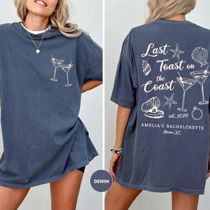 K&ouml;nnte beinhalten: Denimblaues T-Shirt mit einem Frontmotiv aus zwei Martinigl&auml;sern und einem R&uuml;ckenmotiv mit dem Text "Last Toast on the Coast", Muscheln, Seesternen und Martinigl&auml;sern. Der Text "Amelia's Bachelorette Miami, FL" ist ebenfalls auf der R&uuml;ckseite aufgedruckt.