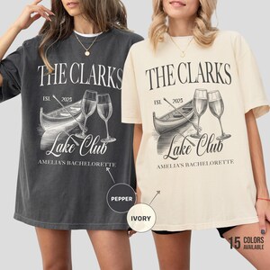 May include: Two oversized t-shirts, one pepper and one ivory, featuring a boat, wine glasses, and the text "THE CLARKS LAKE CLUB AMELIA'S BACHELORETTE EST. 2025". The shirts are casual and comfortable.