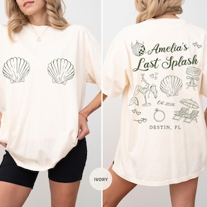 Puede incluir: Camiseta de color marfil con gr&aacute;ficos de conchas marinas en la parte delantera. La parte trasera presenta el texto "Amelia's Last Splash" con ilustraciones de una bola de discoteca, una sirena, champ&aacute;n, un anillo y sillas de playa. Tambi&eacute;n se incluye el texto "Destin, FL".