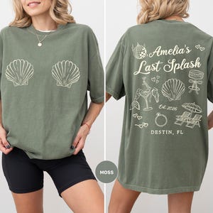 Puede incluir: Camiseta verde musgo con gr&aacute;ficos de conchas blancas en la parte delantera. La parte trasera de la camiseta presenta el texto "Amelia's Last Splash" con gr&aacute;ficos adicionales. La camiseta es de corte holgado.