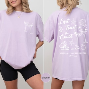 K&ouml;nnte beinhalten: Orchideenfarbenes T-Shirt mit wei&szlig;er Grafik von zwei Martinigl&auml;sern auf der Vorderseite. Auf der R&uuml;ckseite steht der Text "Last Toast on the Coast" mit Muschel- und Ringillustrationen. "Amelia's Bachelorette" ist ebenfalls auf der R&uuml;ckseite aufgedruckt.