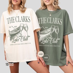 May include: Two t-shirts, one ivory and one moss green, featuring the text "THE CLARKS" above an illustration of a boat and champagne glasses. The text "Lake Club" and "AMELIA'S BACHELORETTE" are also displayed. The shirts are part of a collection with 15 colors available.