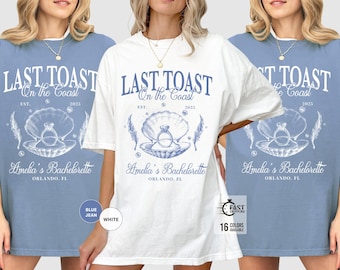 Op de afbeelding: Een wit of blauw spijkerstof T-shirt met de tekst "LAST TOAST On the Coast" en "Amelia's Bachelorette, Orlando, FL" in blauw. Het ontwerp bevat een zeeschelp met een ring, veren en het jaar 2025.