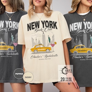Puede incluir: Tres camisetas para mujeres con un estampado gráfico de un taxi amarillo frente al horizonte de la ciudad de Nueva York. El texto "New York Bach Club" está impreso en las camisetas. Las camisetas están disponibles en tres colores: pimienta, marfil y negro.