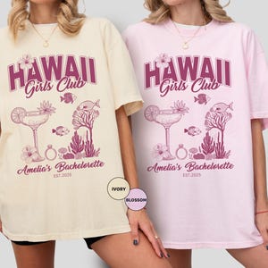 K&ouml;nnte beinhalten: Zwei &uuml;bergro&szlig;e T-Shirts, eines in Elfenbein und eines in Rosa, mit einem lila Aufdruck. Der Aufdruck lautet "HAWAII Girls Club" &uuml;ber einem Cocktail-, Fisch- und Korallen-Design. Darunter steht "Amelia's Bachelorette EST.2025".