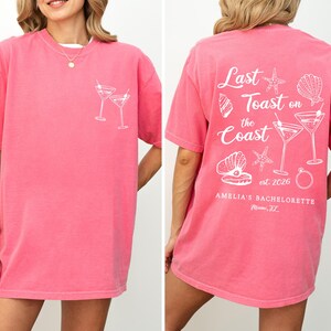 K&ouml;nnte beinhalten: Ein pinkfarbenes T-Shirt mit wei&szlig;en Grafiken. Die Vorderseite zeigt zwei Martinigl&auml;ser, w&auml;hrend die R&uuml;ckseite den Text "Last Toast on the Coast" mit Muschel- und Ringillustrationen zeigt. Das Shirt ist f&uuml;r Amelia's Bachelorette in Miami, FL.