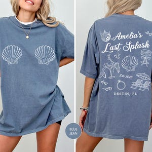Puede incluir: Camiseta azul vaquera con gr&aacute;ficos de conchas blancas en la parte delantera. La parte trasera de la camiseta presenta el texto "Amelia's Last Splash" con ilustraciones de tem&aacute;tica playera y la ubicaci&oacute;n "Destin, FL".